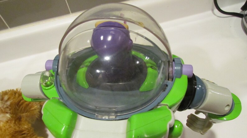 May include: A close-up of a Buzz Lightyear action figure. The toy has a clear plastic helmet, a purple head, and a green and white body. The figure is a popular character from the Toy Story franchise.