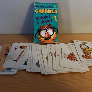 May include: A Garfield Riddles & Jokes card game set. The box is teal with Garfield's face and the text "Riddles & Jokes #1". Several cards are fanned out, each with Garfield illustrations and text.