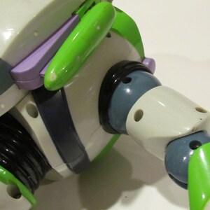 May include: Close-up of a Buzz Lightyear action figure. The toy features a white body with green and purple accents. The figure's arm is articulated, with a black joint and a green wing-like detail. The figure has a gray helmet and a black neck.