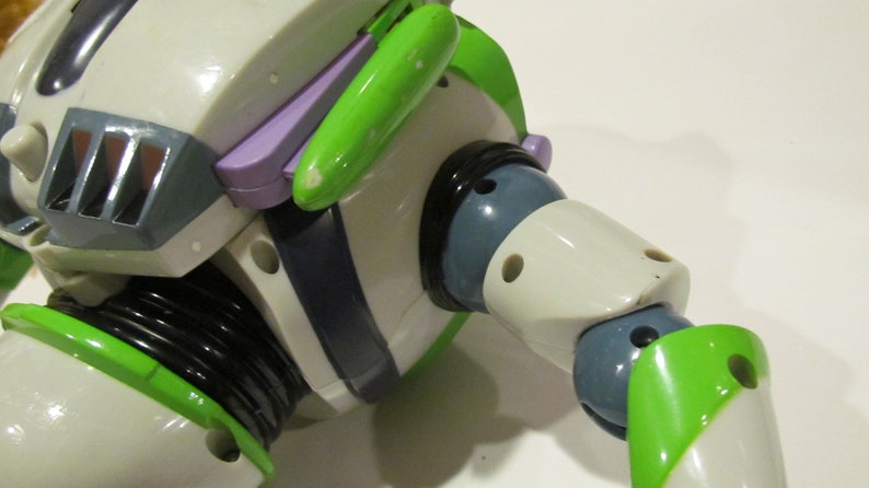 May include: Close-up of a Buzz Lightyear action figure. The toy features a white body with green and purple accents. The figure's arm is articulated, with a black joint and a green wing-like detail. The figure has a gray helmet and a black neck.