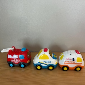 May include: Three toy emergency vehicles: a red fire truck with "Fire Truck" text, a white and blue police car with "Police" text, and a white and orange ambulance with an emblem. Each has black wheels.