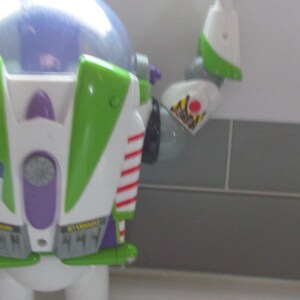 Puede incluir: Primer plano de una figura de acci&oacute;n de Buzz Lightyear. El juguete es principalmente blanco, verde y morado, con rayas rojas y una c&uacute;pula transparente. El brazo est&aacute; extendido, mostrando una etiqueta de advertencia.