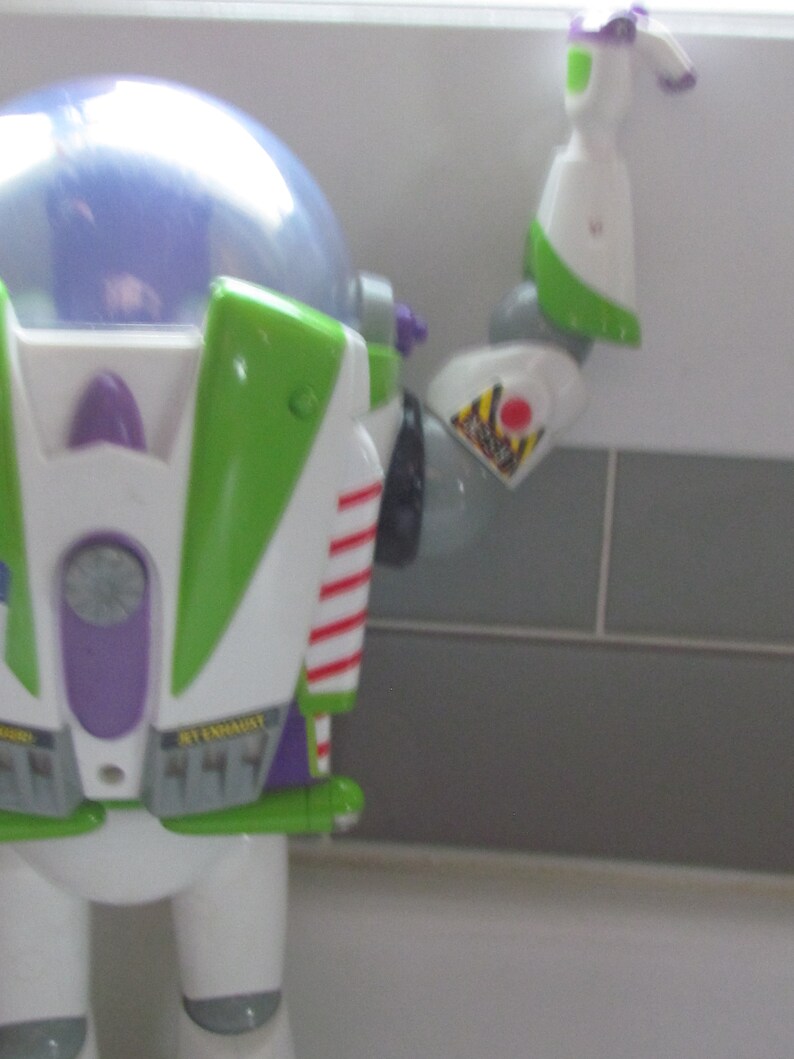Puede incluir: Primer plano de una figura de acci&oacute;n de Buzz Lightyear. El juguete es principalmente blanco, verde y morado, con rayas rojas y una c&uacute;pula transparente. El brazo est&aacute; extendido, mostrando una etiqueta de advertencia.