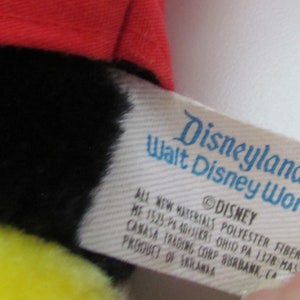 May include: A plush toy with a red hat, black fur, and yellow accents. The tag reads "Disneyland Walt Disney World" and includes the Disney copyright. The toy is made of polyester fibers.