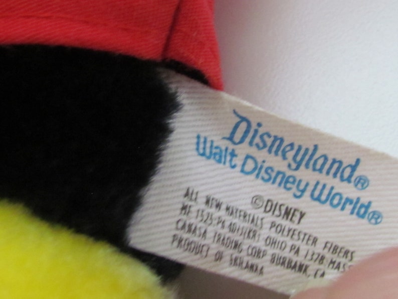 May include: A plush toy with a red hat, black fur, and yellow accents. The tag reads "Disneyland Walt Disney World" and includes the Disney copyright. The toy is made of polyester fibers.