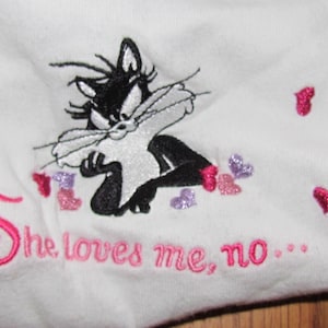 May include: White fabric with an embroidered cartoon cat in black and white, with a sassy expression. Pink and purple glitter hearts are scattered around the cat. The text "She loves me, no..." is embroidered in pink.