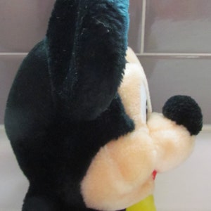 May include: A plush toy of a cartoon mouse, featuring a black head and ears, a cream-colored face, and a black nose. The toy has a yellow shirt and a red mouth. The plush is soft and fluffy.