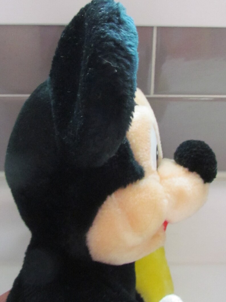 May include: A plush toy of a cartoon mouse, featuring a black head and ears, a cream-colored face, and a black nose. The toy has a yellow shirt and a red mouth. The plush is soft and fluffy.
