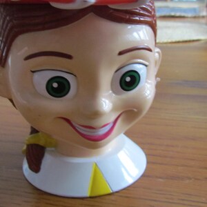 May include: A plastic toy cup shaped like the head of a girl with brown hair, green eyes, and a red and white hat. The cup has a yellow and white collar.