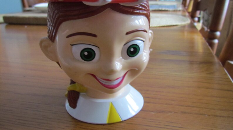 May include: A plastic toy cup shaped like the head of a girl with brown hair, green eyes, and a red and white hat. The cup has a yellow and white collar.