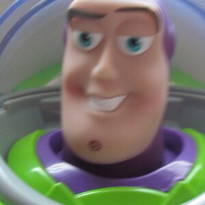 Puede incluir: Primer plano de una figura de acci&oacute;n de Buzz Lightyear. El juguete tiene un tono de piel claro, una expresi&oacute;n sonriente y ojos azules. La figura lleva un traje espacial verde y morado con un casco transparente.