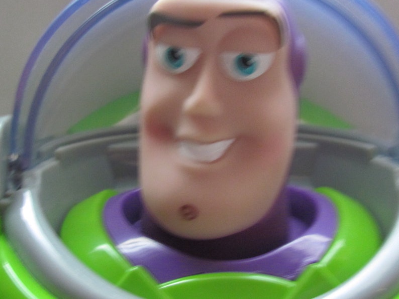 Puede incluir: Primer plano de una figura de acci&oacute;n de Buzz Lightyear. El juguete tiene un tono de piel claro, una expresi&oacute;n sonriente y ojos azules. La figura lleva un traje espacial verde y morado con un casco transparente.