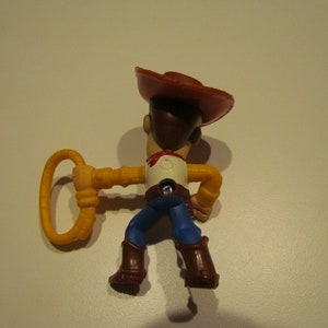 May include: A plastic toy figure of Woody from Toy Story. Woody is wearing his signature brown cowboy hat, blue jeans, and a yellow shirt. He is holding a yellow lasso in his right hand.