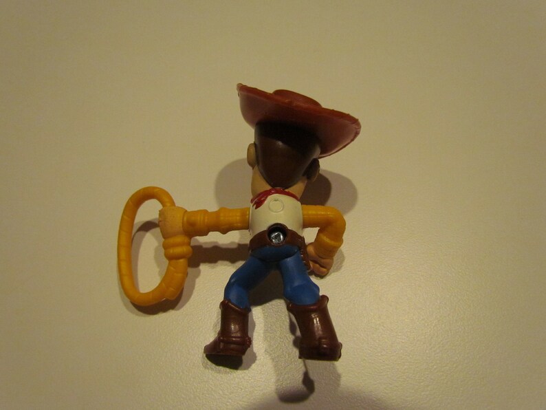 May include: A plastic toy figure of Woody from Toy Story. Woody is wearing his signature brown cowboy hat, blue jeans, and a yellow shirt. He is holding a yellow lasso in his right hand.