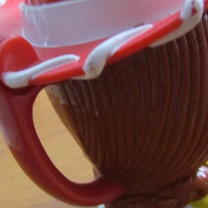May include: Red and white plastic mug with a handle and a cartoon character design. The mug is shaped like a cowboy hat.