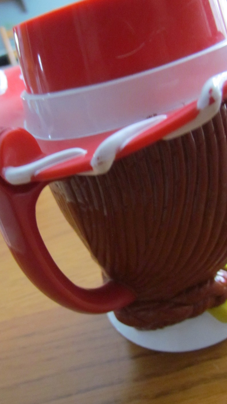 May include: Red and white plastic mug with a handle and a cartoon character design. The mug is shaped like a cowboy hat.