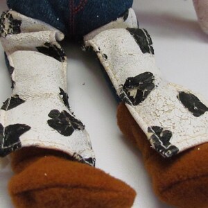 Pode incluir: Close-up das pernas e botas de uma boneca de brinquedo. As botas s&atilde;o castanhas com polainas brancas com manchas pretas. As polainas t&ecirc;m um aspeto envelhecido e rachado. Cal&ccedil;as de ganga azuis s&atilde;o vis&iacute;veis acima das botas.