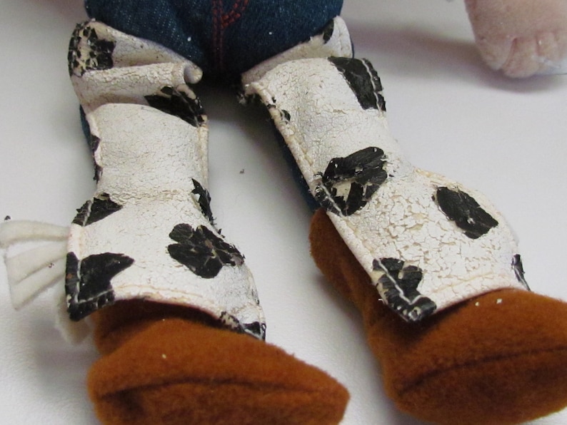 Pode incluir: Close-up das pernas e botas de uma boneca de brinquedo. As botas s&atilde;o castanhas com polainas brancas com manchas pretas. As polainas t&ecirc;m um aspeto envelhecido e rachado. Cal&ccedil;as de ganga azuis s&atilde;o vis&iacute;veis acima das botas.