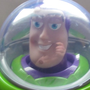 Puede incluir: Primer plano de una figura de acci&oacute;n de Buzz Lightyear. El juguete tiene un traje espacial verde y morado con un casco transparente. La cara de la figura es de color rosa claro con ojos azules y una ligera sonrisa. El tema general son los juguetes y objetos de colecci&oacute;n.