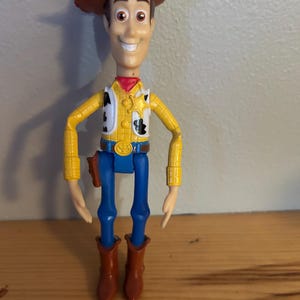 May include: A plastic Woody action figure from the Toy Story franchise. The figure wears a brown cowboy hat, yellow shirt with black cow spots, blue pants, and brown boots. The figure has a red neckerchief and a sheriff's badge.