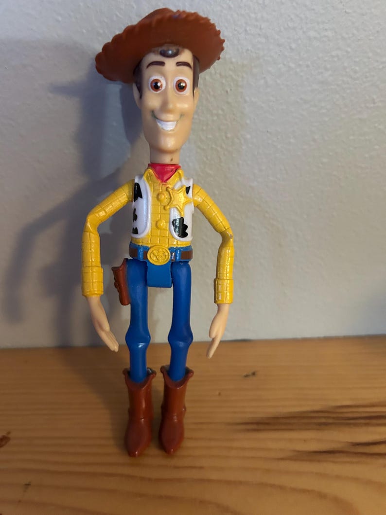 May include: A plastic Woody action figure from the Toy Story franchise. The figure wears a brown cowboy hat, yellow shirt with black cow spots, blue pants, and brown boots. The figure has a red neckerchief and a sheriff's badge.