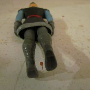 K&ouml;nnte beinhalten: Eine Kunststoff-Actionfigur eines Charakters in dunkler Kleidung mit rotem G&uuml;rtel, blauen &Auml;rmeln und grauen Stiefeln. Die Figur liegt auf einer hellen Oberfl&auml;che.