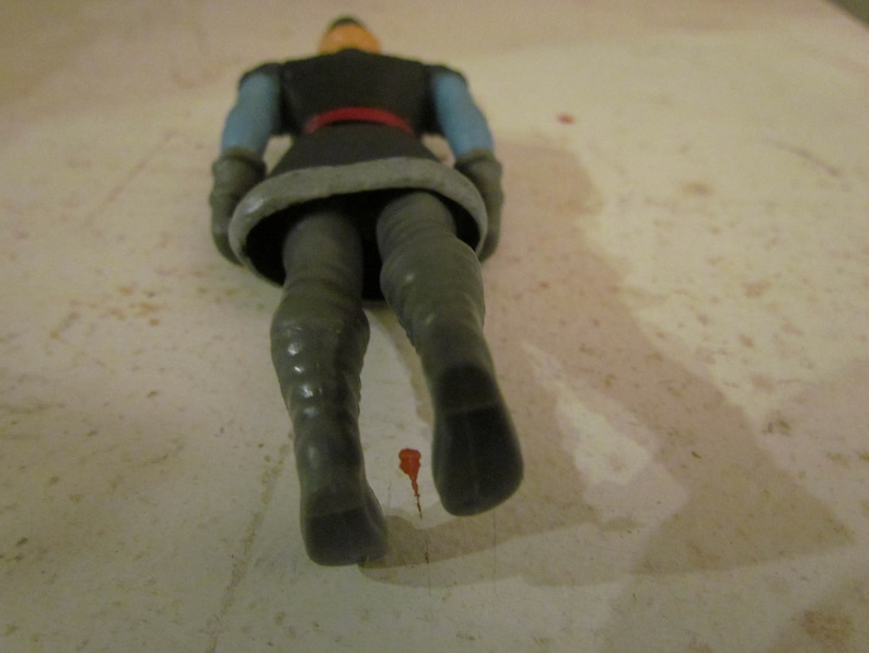 K&ouml;nnte beinhalten: Eine Kunststoff-Actionfigur eines Charakters in dunkler Kleidung mit rotem G&uuml;rtel, blauen &Auml;rmeln und grauen Stiefeln. Die Figur liegt auf einer hellen Oberfl&auml;che.