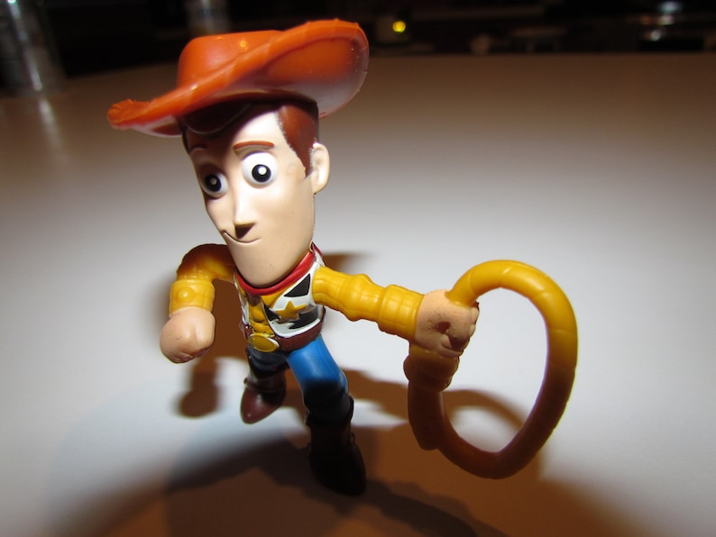May include: A plastic toy figurine of Woody from Toy Story. Woody is wearing his signature brown cowboy hat, a yellow vest, and blue jeans. He is holding a yellow lasso in his right hand.