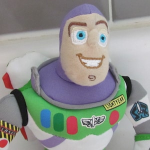May include: A plush toy of Buzz Lightyear from the Toy Story franchise. The toy is wearing his signature space ranger suit with a purple helmet, green vest, and white undershirt. The toy has a friendly face with blue eyes and a big smile. The toy's chest features a patch that says "LIGHTYEAR".