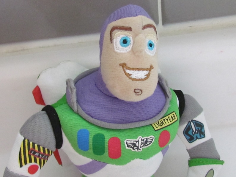 May include: A plush toy of Buzz Lightyear from the Toy Story franchise. The toy is wearing his signature space ranger suit with a purple helmet, green vest, and white undershirt. The toy has a friendly face with blue eyes and a big smile. The toy's chest features a patch that says "LIGHTYEAR".