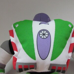 May include: A green, white, and purple plush toy of Buzz Lightyear from the Toy Story franchise. The toy is facing away from the camera, showing the back of the space ranger's suit.