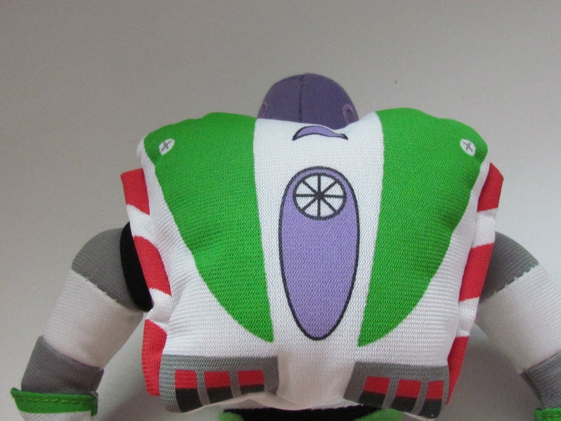 May include: A green, white, and purple plush toy of Buzz Lightyear from the Toy Story franchise. The toy is facing away from the camera, showing the back of the space ranger's suit.