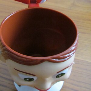 May include: A brown and white plastic mug shaped like a person's head. The mug has a brown rim and a white face with brown hair and green eyes.