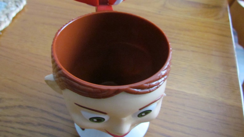 May include: A brown and white plastic mug shaped like a person's head. The mug has a brown rim and a white face with brown hair and green eyes.
