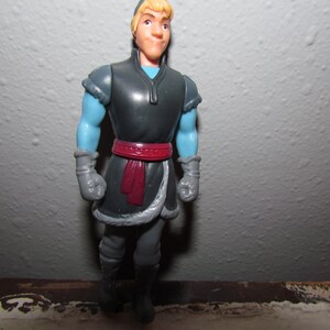 K&ouml;nnte beinhalten: Eine Plastikfigur einer Figur aus dem Animationsfilm "Die Eisk&ouml;nigin". Die Figur hat blondes Haar, blaue Arme und tr&auml;gt ein dunkelgraues Outfit mit rotem G&uuml;rtel und grauen Handschuhen. Die Figur steht aufrecht.
