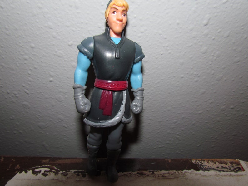 K&ouml;nnte beinhalten: Eine Plastikfigur einer Figur aus dem Animationsfilm "Die Eisk&ouml;nigin". Die Figur hat blondes Haar, blaue Arme und tr&auml;gt ein dunkelgraues Outfit mit rotem G&uuml;rtel und grauen Handschuhen. Die Figur steht aufrecht.