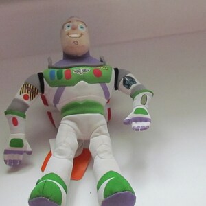 May include: A plush toy of Buzz Lightyear from the Toy Story franchise. The toy is wearing his signature space ranger suit with green, white, and purple accents. He has a friendly face with blue eyes and a big smile.