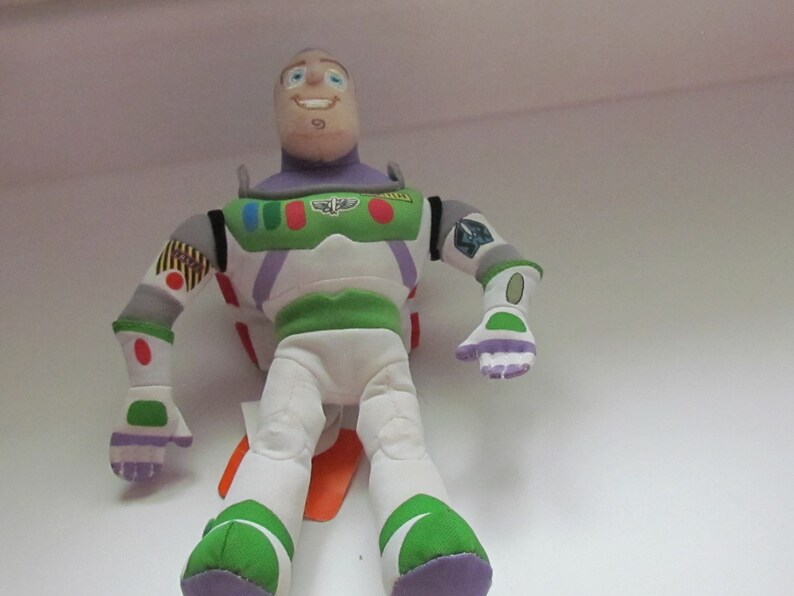 May include: A plush toy of Buzz Lightyear from the Toy Story franchise. The toy is wearing his signature space ranger suit with green, white, and purple accents. He has a friendly face with blue eyes and a big smile.