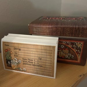 May include: A rectangular address box with a wood grain finish and the word ADDRESSES printed on top. Beside it sits a stack of white address cards featuring a floral illustration of tulips and daisies, with printed lines for contact details.