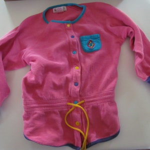 May include: A pink corduroy romper with a blue and yellow accent. The romper has a blue collar and cuffs, a small pocket with a diamond design, and a yellow drawstring. Buttons are yellow and blue.