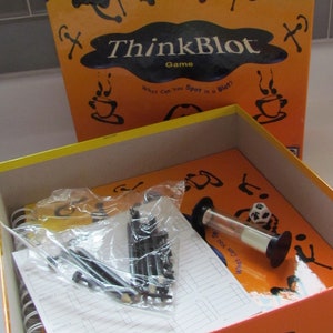 May include: The ThinkBlot game box is open, revealing the game's components: a spiral-bound booklet, a sand timer, a die, and black game pieces. The box is orange with black and white text.