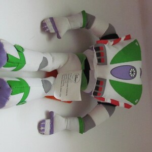 May include: A green, white, and purple plush toy of Buzz Lightyear from the Toy Story franchise. The toy is lying on its back with its arms and legs outstretched. The toy has a tag attached to it that reads "Disney".