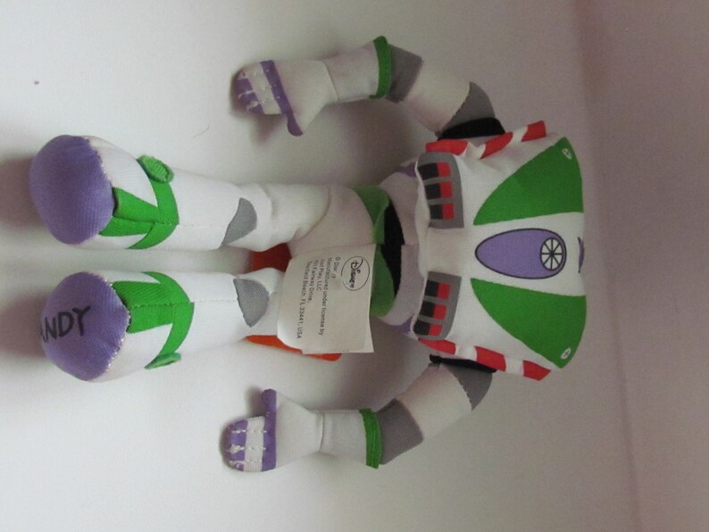 May include: A green, white, and purple plush toy of Buzz Lightyear from the Toy Story franchise. The toy is lying on its back with its arms and legs outstretched. The toy has a tag attached to it that reads "Disney".