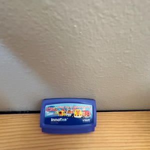 May include: Blue plastic cartridge for the InnoTab learning tablet. The cartridge features a colorful image of a bus, a police car, a boat, and a fire truck. The text on the cartridge reads "Busy Day in Alphabet Town".
