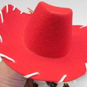 Pode incluir: Um chap&eacute;u de cowboy vermelho brilhante com uma coroa arredondada e uma aba larga. A aba &eacute; acentuada com costuras brancas. Um acess&oacute;rio cl&aacute;ssico do oeste.