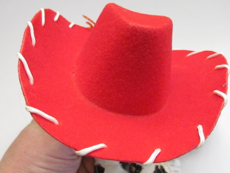 Pode incluir: Um chap&eacute;u de cowboy vermelho brilhante com uma coroa arredondada e uma aba larga. A aba &eacute; acentuada com costuras brancas. Um acess&oacute;rio cl&aacute;ssico do oeste.