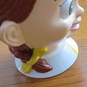 May include: A brown and white ceramic figurine of a girl with brown hair and a yellow bow. The figurine is smiling and has green eyes.