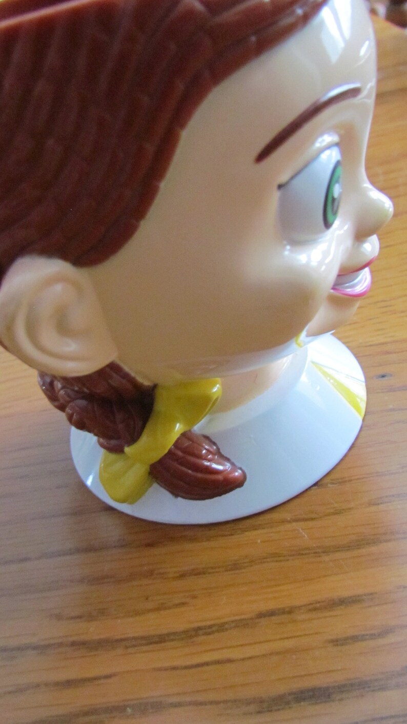 May include: A brown and white ceramic figurine of a girl with brown hair and a yellow bow. The figurine is smiling and has green eyes.