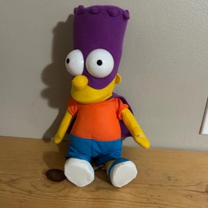 May include: A plush Bart Simpson doll wearing a purple hat, cape, and blue shorts. The doll has large, white eyes, a yellow face, and an orange shirt. The doll is sitting on a wooden surface.