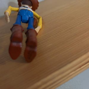 May include: A Woody action figure, viewed from the back, with a brown cowboy hat, yellow shirt, blue jeans, and brown boots. The figure is positioned on a wooden surface, showcasing the character's iconic outfit.